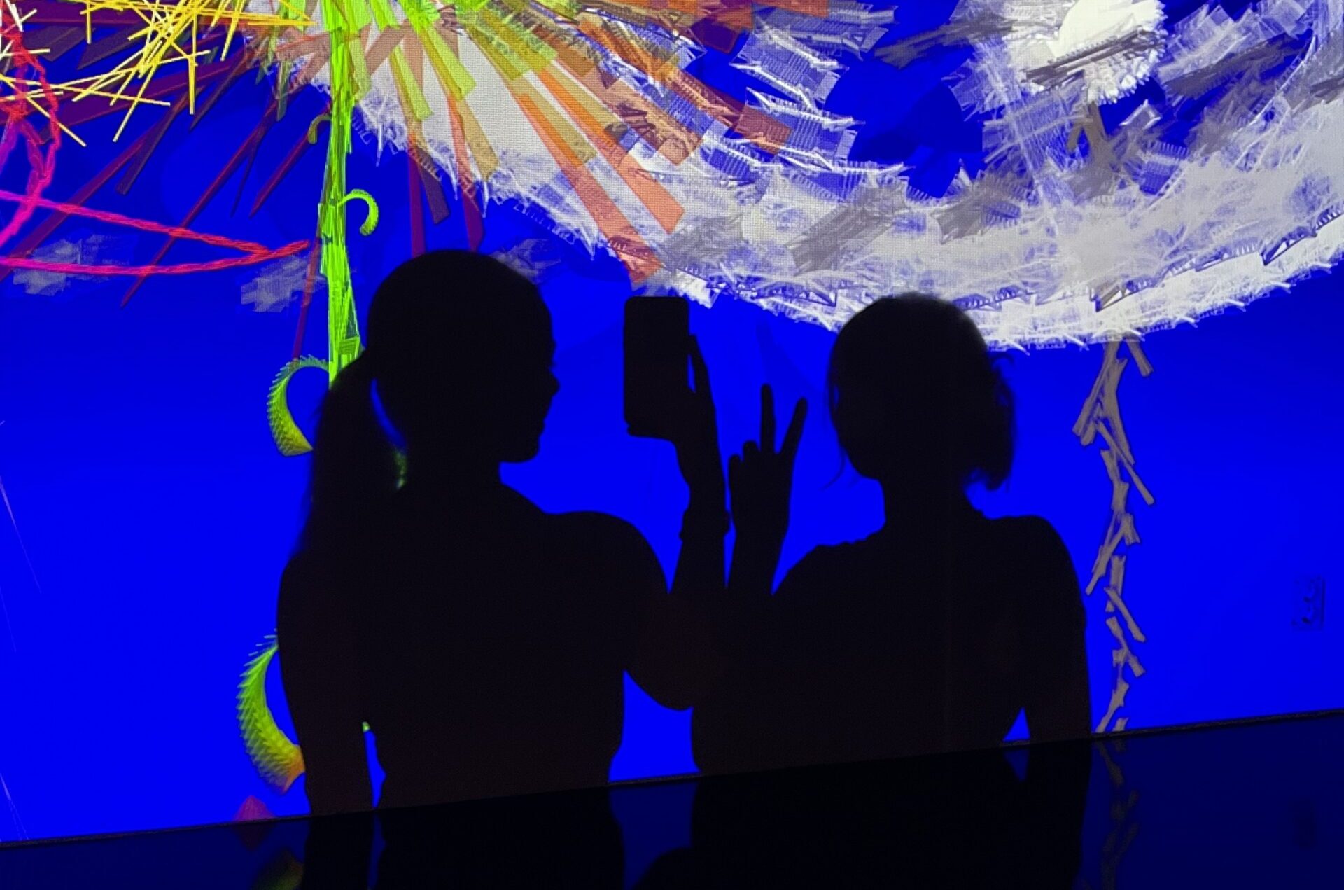 Two shadows against blue light show wall, one girl doing peace sign